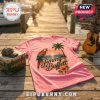 Pink crew neck t-shirt featuring a parrot, palm tree, sunset, and Jimmy Buffett script logo on a wooden dock background.!