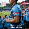 Man wearing a light blue NSW Blues rugby polo shirt featuring player portraits and team branding in a crowded stadium.!