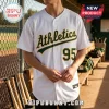 White Athletics baseball jersey with green and yellow text and number 95 on front, worn by a person in a baseball dugout.!