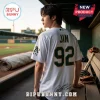 Jin from BTS wearing a white Oakland Athletics-style baseball jersey with number 92 and custom branding in a dugout.!