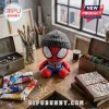 Spider-Man Brand New Day Beanie Plush Toy wearing a gray knit hat, sitting on a wooden desk with art supplies.