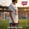 Man wearing a white USA soccer jersey with blue star sleeves and red stripes, holding brown soccer cleats on a field.!
