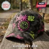 Blink-182 Neon Smiley Camo Cap with pink and green accents sitting on a weathered wooden park bench.
