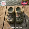 The Walking Dead Olive Green Clogs with Charms featuring miniature post-apocalyptic weapon and gear icons on a wood dock.
