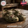 The Walking Dead Olive Green Clogs with Charms featuring miniature sheriff hat, katana, and series logo on wooden table.