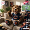 Front view of a brown plush bear mascot sitting on a couch wearing a blue jersey and pointing upward.