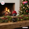 Close-up view of a dark puppy wearing a red knit hat while carrying firewood, set before a fireplace and Christmas tree.
