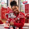 Smiling woman hugging a crochet elephant plush doll wearing a hat and sweater with Greek letters.