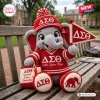 Crochet elephant plush doll wearing a red sweater and hat with Greek letters on a wooden bench.