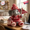 Front view of a crochet elephant plush doll wearing a red hat, sweater, and holding a flag with Greek letters.