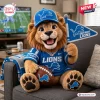 Front view of a plush lion wearing a team jersey and hat while holding a pennant flag and football on a grey couch.