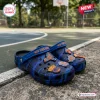 Side view of New York Knicks Blue Camo Clogs with Charms resting on an outdoor basketball court.