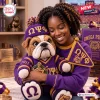 A woman hugging an Omega Psi Phi bulldog crochet plush toy wearing a matching purple and gold knit hat and sweater.