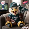 Front view of an eagle mascot plush doll wearing a jersey and cap, holding a flag and football on a gray couch.