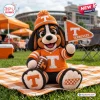 Front view of a crocheted mascot plush holding a football and a pennant on a plaid blanket at a stadium.