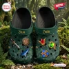 Top view of green clog shoes featuring decorative charms and Zelda branding resting on mossy ground.