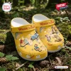 Top view of yellow Winnie the Pooh clogs with character charms resting on forest ground.