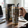 Hand holding an Alpha Phi Alpha Sphinx Stainless Steel Tumbler with gold trim and matching gift box on a desk.