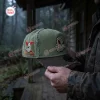 Side view of an olive green Anaheim Ducks x Jason Voorhees baseball cap held outdoors near a cabin.