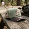 Side angle of an olive green baseball cap with a brown brim and hockey mask logo resting on a weathered wooden picnic table.