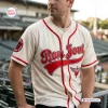 Front view of a man wearing a cream baseball jersey with red piping, red lettering, and a winged heart emblem in a stadium.