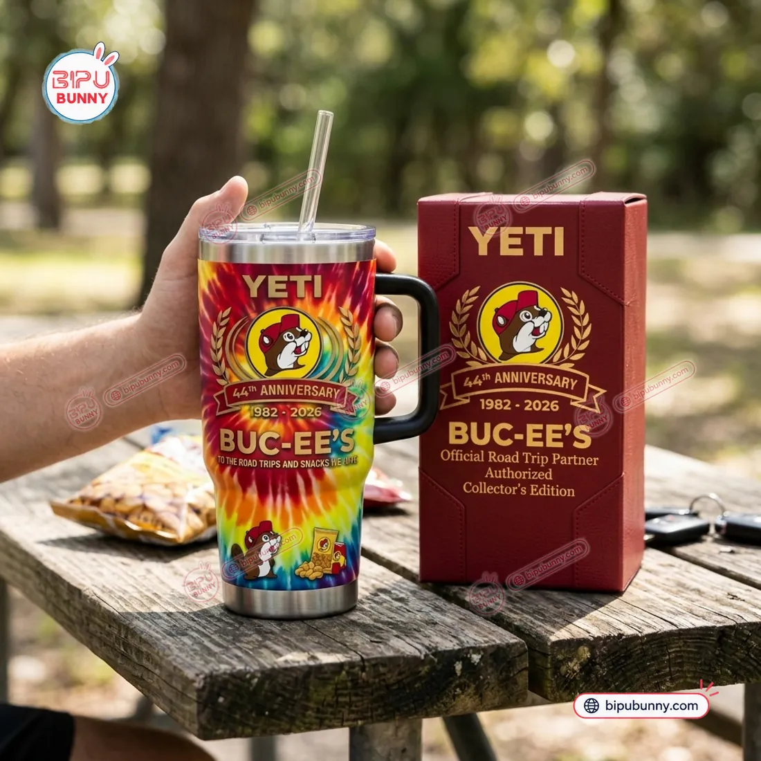 Buc-ees 44th Anniversary Tumbler Cup held in hand on a wooden picnic table next to its maroon collector box