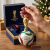 Closeup of hands holding a FIFA World Cup 2026 Golden Trophy & Trionda Ball Christmas Ornament above its blue box.