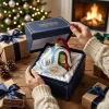 Hands opening a blue gift box containing a Trionda Ball Christmas Ornament in a holiday setting.
