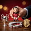 Close-up of a hand holding a Manchester United figure next to a miniature trophy and Old Trafford stadium.