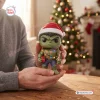 Close-up view of hands holding a green figure wearing a Santa hat and holding a gift in front of a tree.