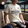 Front view of a cream baseball jersey with World Series Champions text worn by a seated person in a dugout.