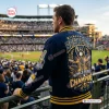 Rear view of a man wearing a navy blue varsity jacket with embroidered graphics inside a baseball stadium.