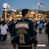 Back view of a fan wearing a Milwaukee Brewers 2026 World Series Championship Baseball Jacket at a lit stadium.