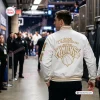 Rear view of a man wearing a white New York Knicks 2026 Champions baseball jacket with gold embroidery in an arena hallway.