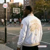 Rear view of a man wearing a white New York Knicks 2026 Champions Baseball Jacket with gold lettering at an outdoor basketball court.