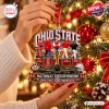 Close-up of hands holding an Ohio State Buckeyes National Champions Christmas ornament in front of a lit tree.