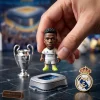 Closeup of a hand holding a miniature soccer figurine wearing a white jersey next to a trophy replica and crest pin.