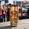 Front view of a San Francisco 49ers 80th Anniversary tumbler cup with gold finish, red handle, and lid outdoors.
