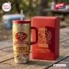 Front view of the San Francisco 49ers Gold 80th Anniversary Tumbler with straw next to a red box on a wooden table.