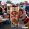 Hand holding a San Francisco 49ers Gold 80th Anniversary Tumbler with handle outdoors.