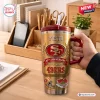 Hand holding a San Francisco 49ers Gold 80th Anniversary Tumbler with handle on a wooden desk.
